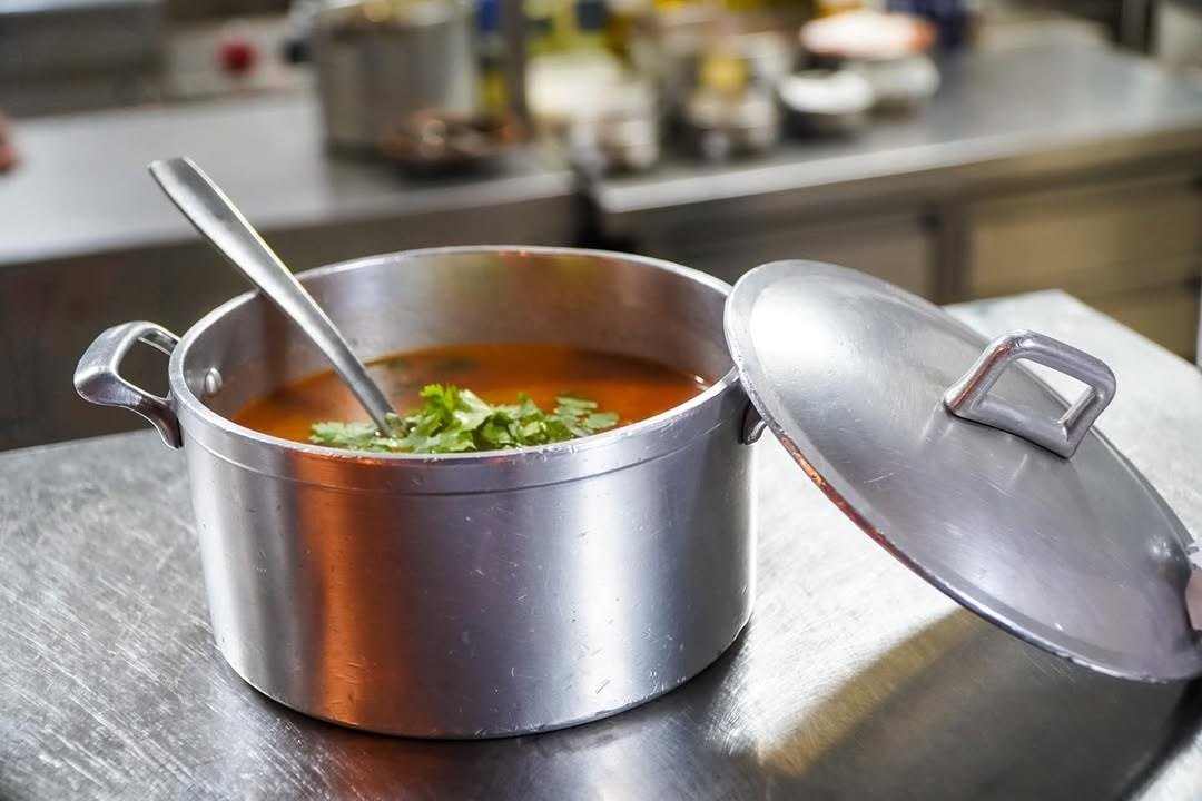 Panela de alumínio com sopa e ervas frescas, de tampa aberta, numa bancada de cozinha em inox